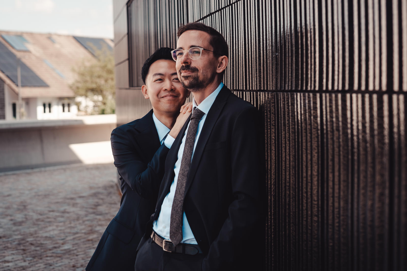 Hochzeitsfotograf München | Hochzeitsfotograf München Two businessmen in suits leaning against a dark striped wall, one smiling as the other adjusts his tie behind him. - Hochzeitsfotograf München