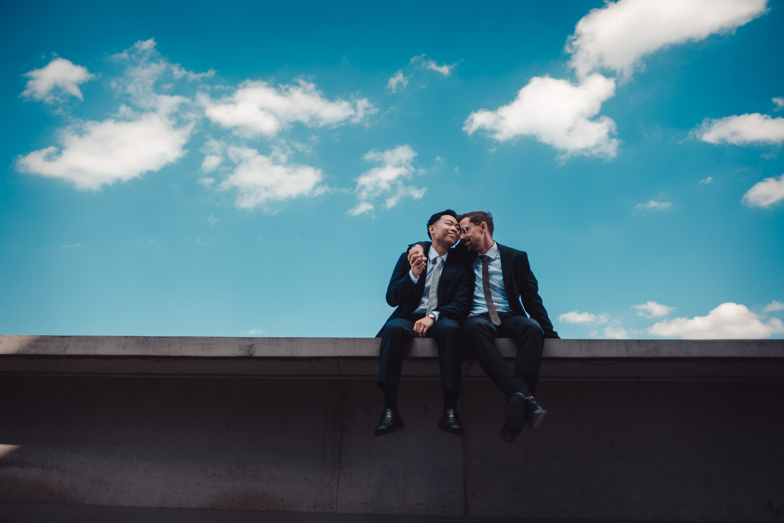 Hochzeitsfotograf München | Hochzeitsfotograf München Two men in dark suits sit close together on a rooftop ledge, leaning in and smiling at each other against a bright blue sky with scattered clouds. - Hochzeitsfotograf München