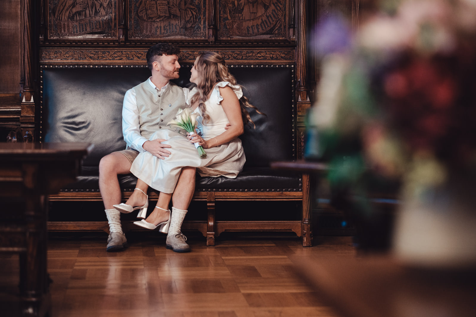 Hochzeitsfotograf München | Hochzeitsfotograf München Bride and groom in wedding attire sit close on a carved wooden bench, gazing at each other; the bride holds a bouquet of white lilies. - Hochzeitsfotograf München