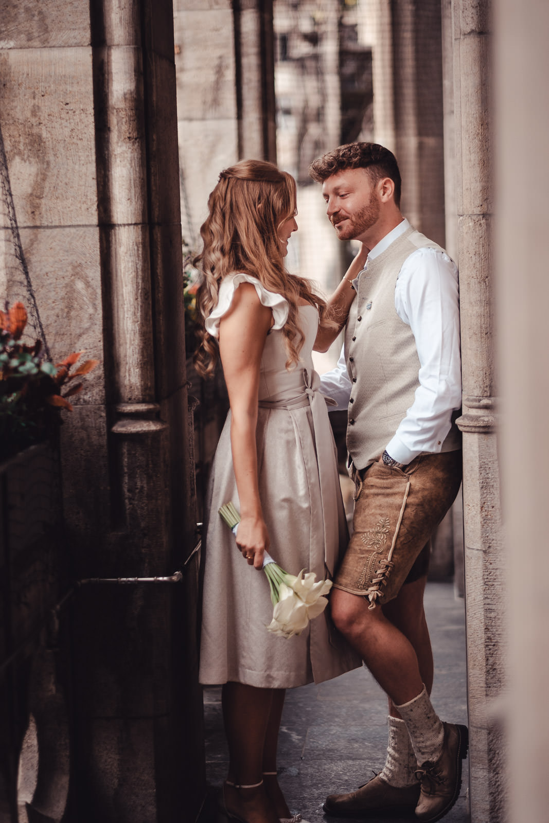 Hochzeitsfotograf München | Hochzeitsfotograf München Romantic moment between a couple in a narrow stone alley; woman in a beige dress holds white flowers, man in a beige vest smiles at her. - Hochzeitsfotograf München