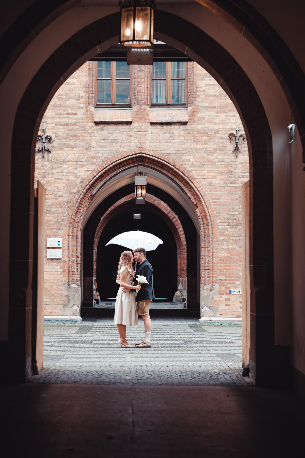 Hochzeitsfotograf München | Hochzeitsfotograf München Couple sharing a kiss under a brick archway, woman in a light dress holding a bouquet, man in a blazer and shorts, white umbrella above them. - Hochzeitsfotograf München