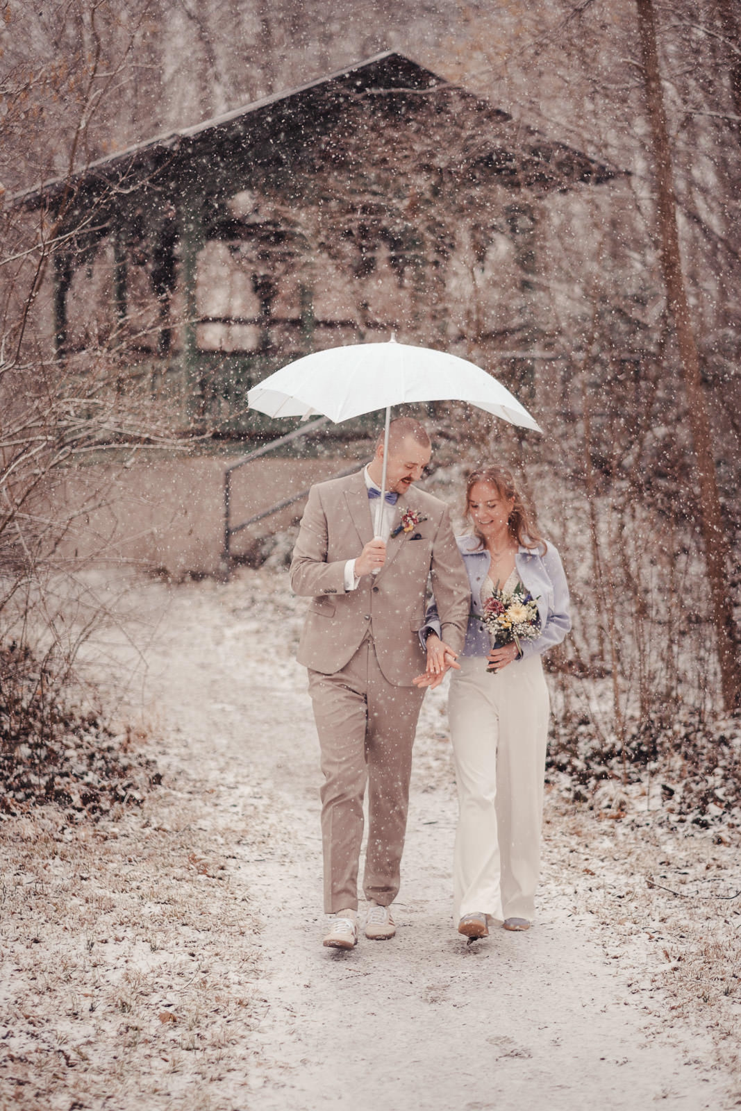 Hochzeitsfotograf München | Hochzeitsfotograf München Couple walking hand in hand in a snowy park, sharing a white umbrella during a winter wedding moment with a rustic cabin in the background. - Hochzeitsfotograf München