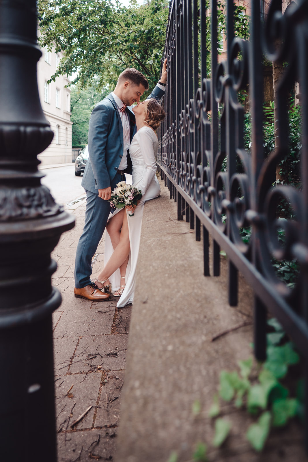 Hochzeitsfotograf München | Hochzeitsfotograf München Bride and groom sharing a kiss beside a wrought-iron fence on an urban sidewalk; she holds a bouquet in a white dress. - Hochzeitsfotograf München