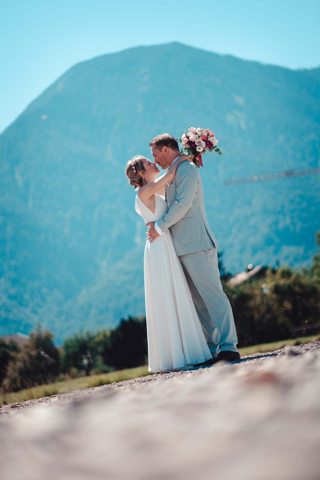 Hochzeitsfotograf München | Hochzeitsfotograf München Bride and groom embrace and kiss outdoors, bouquet in hand, with a blue mountain backdrop behind them. - Hochzeitsfotograf München