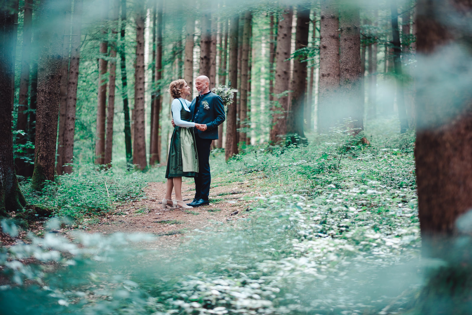 Hochzeitsfotograf München | Hochzeitsfotograf München Couple in a forest path holding hands and smiling at each other among tall trees, bouquet nearby in a serene woodland setting - Hochzeitsfotograf München