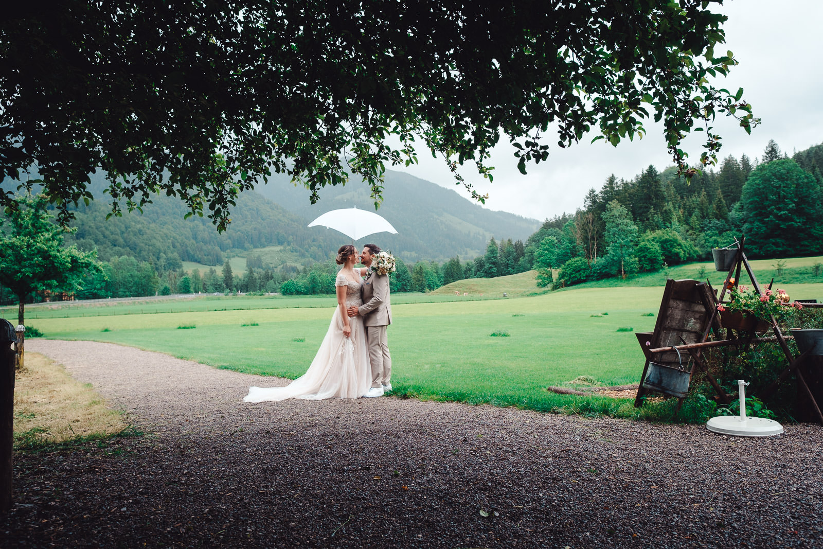Hochzeitsfotograf München | Hochzeitsfotograf München Bride and groom kiss under a white umbrella in a green field with mountains in the background. - Hochzeitsfotograf München