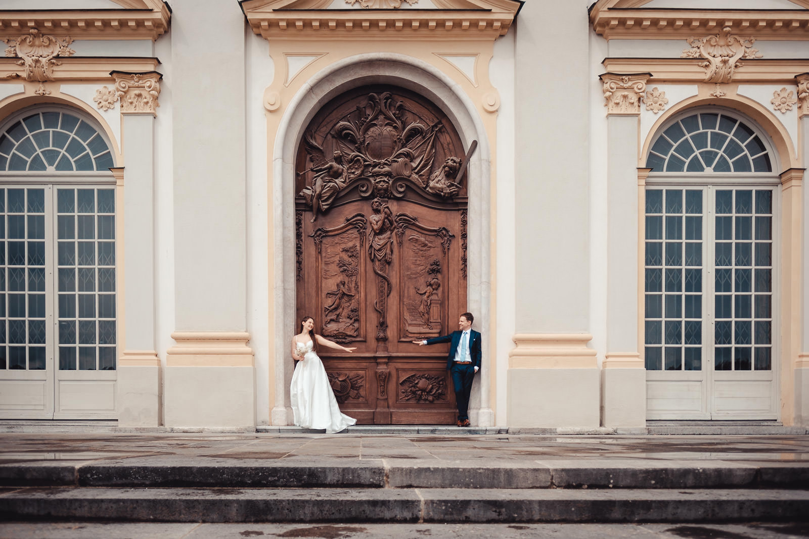 Hochzeitsfotograf München | Hochzeitsfotograf München Bride in a white wedding gown and groom in a suit gesture toward each other in front of an ornate arched doorway on a pale historic building. - Hochzeitsfotograf München