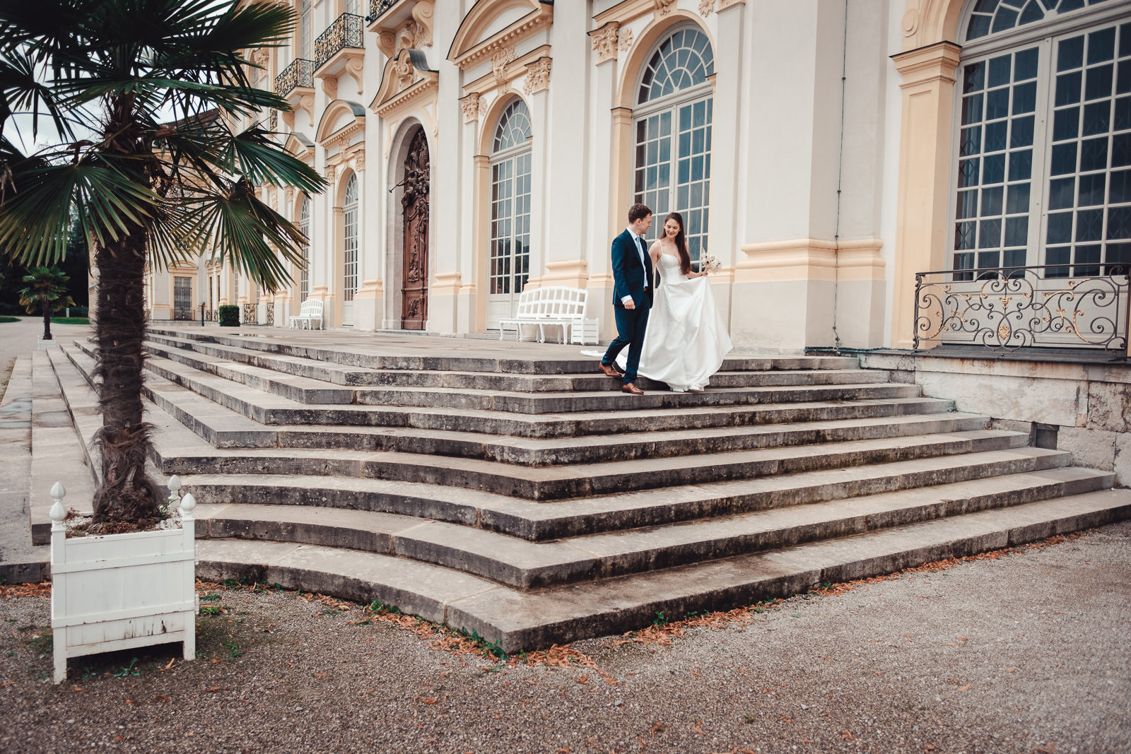 Hochzeitsfotograf München | Hochzeitsfotograf München Bride in a white wedding dress and a groom in a dark suit descend wide stone steps outside a cream neoclassical building with arched windows and ornate details. - Hochzeitsfotograf München