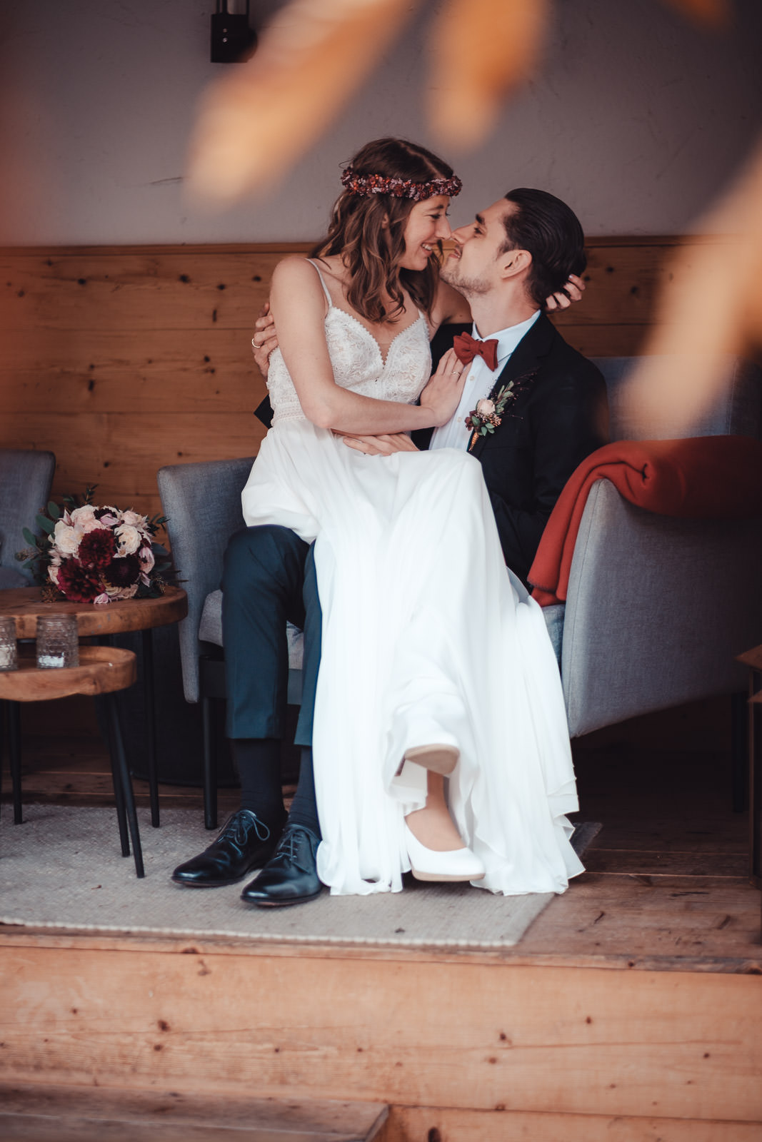 Hochzeitsfotograf München | Hochzeitsfotograf München Bride in a lace wedding dress with a flower crown sits on the groom's lap, smiling as they lean in for a kiss. - Hochzeitsfotograf München