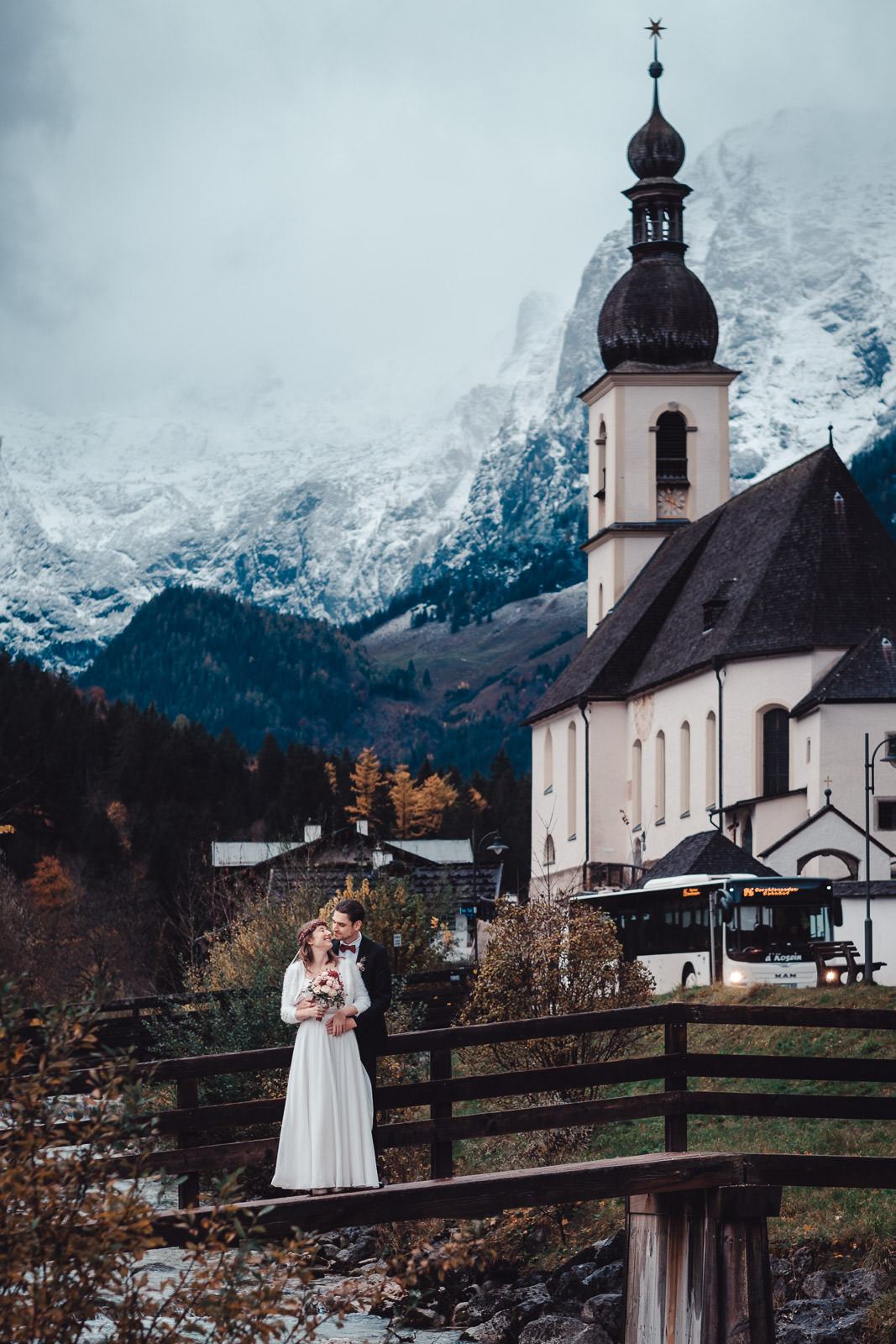 Hochzeitsfotograf München | Hochzeitsfotograf München Bride and groom in wedding attire embrace on a wooden bridge beside a church with snowy mountains in the background. - Hochzeitsfotograf München