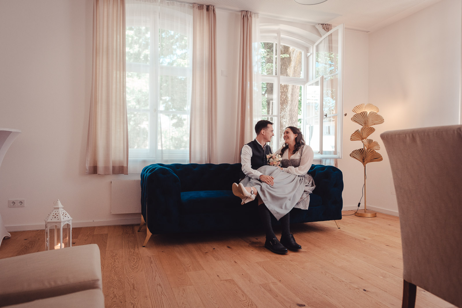 Hochzeitsfotograf München | Hochzeitsfotograf München Couple in vintage attire sits closely on a dark blue velvet sofa in a sunlit living room, smiling at each other with a bouquet in hand. - Hochzeitsfotograf München